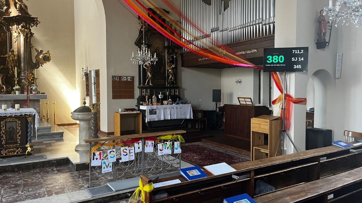 Kirchenraum mit Fernseher an der Wand, der die aktuelle Liednummer 380 groß und die kommenden Nummern daneben anzeigt