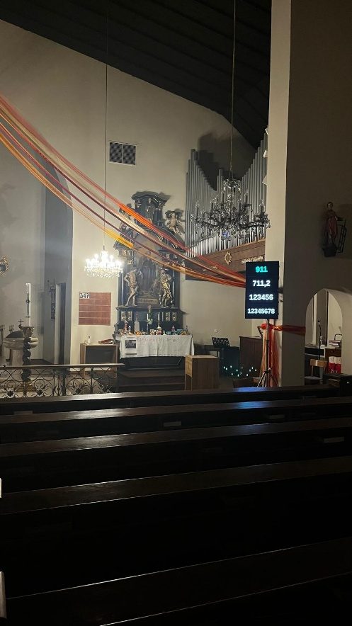 Blick von den Kirchenbänken zum Altar, der Fernseher hängt seitlich am Chorbogen